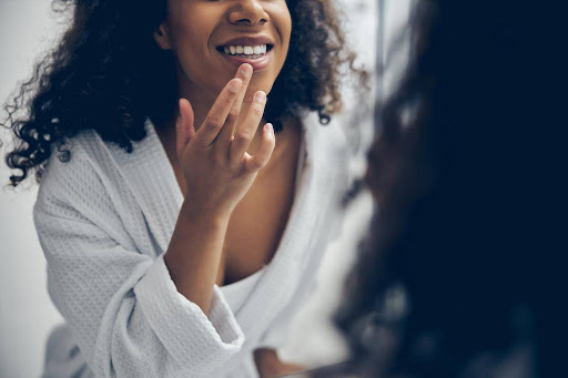 A woman smiling and looking at her teeth in a bathroom mirror. She has one finger pressed against her bottom lip.