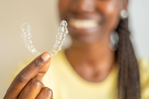 A woman holding up a dental aligner. The camera is focused on the aligner while you can see the blurred image of the smiling woman in the background. 