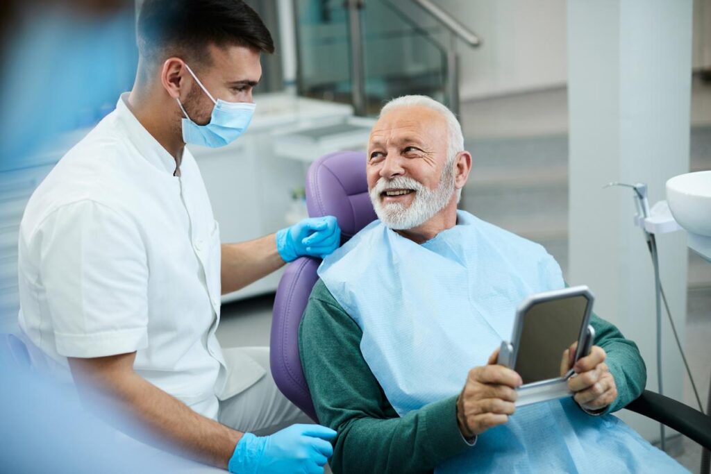 Older man sitting in a dental chair holding a small mirror and talking to the dentist