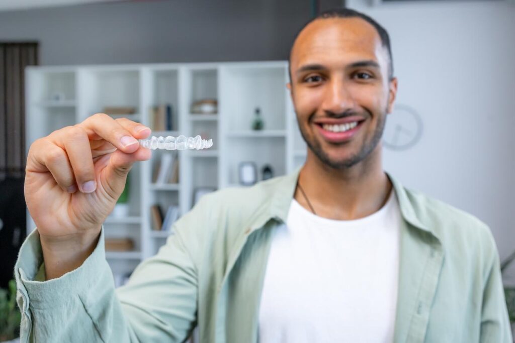 A man wearing a white t-shirt and open, light green button-up shirt smiling and holding up a clear dental aligner tray.
