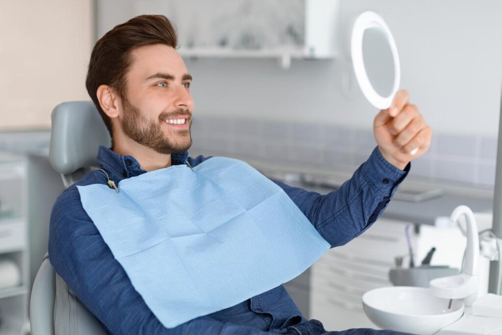 Male patient sitting in a dental chair smiling at his reflection as he looks at his smile in a hand-held mirror.