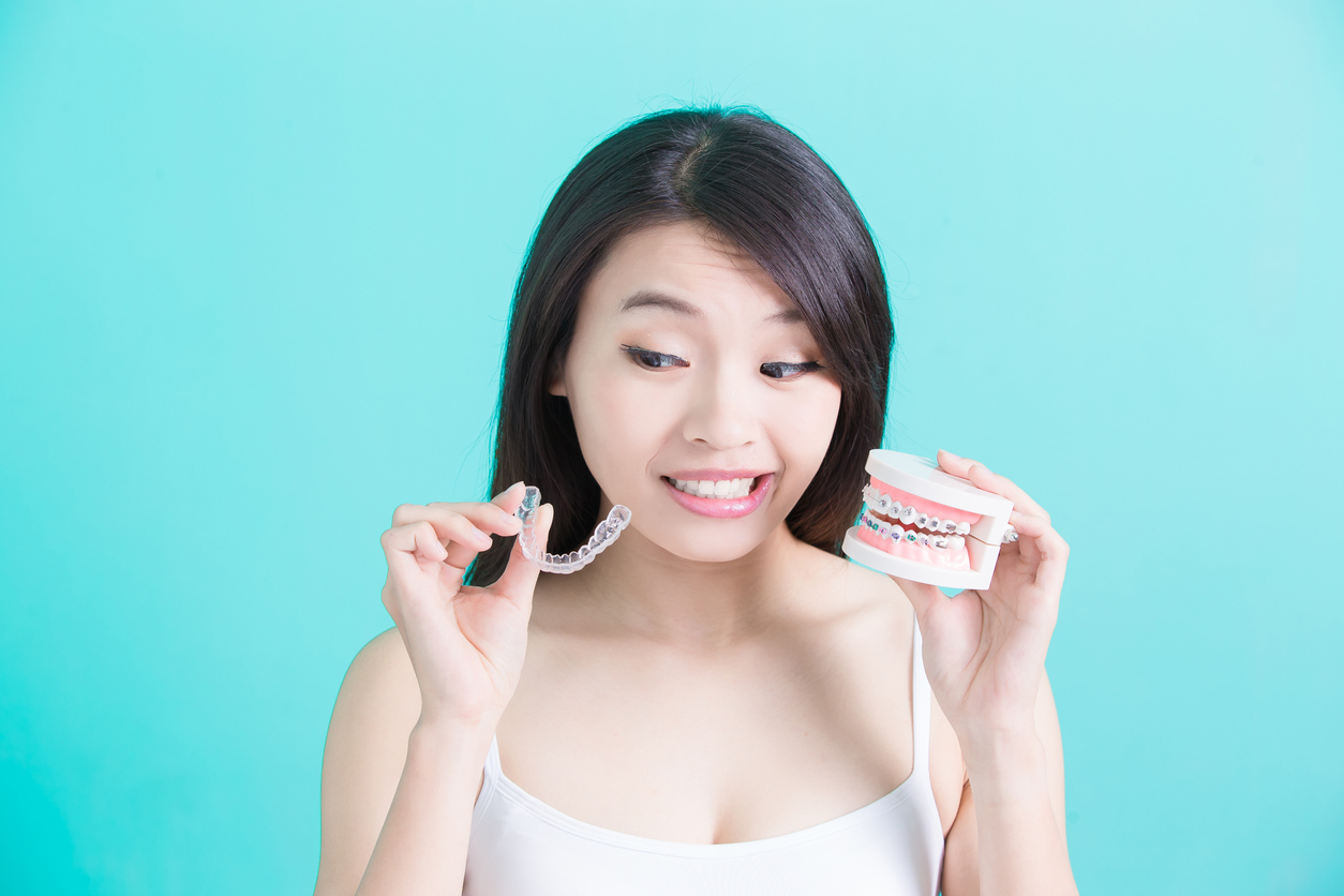 A woman holding up a clear aligner tray like Invisalign and a model of teeth wearing braces